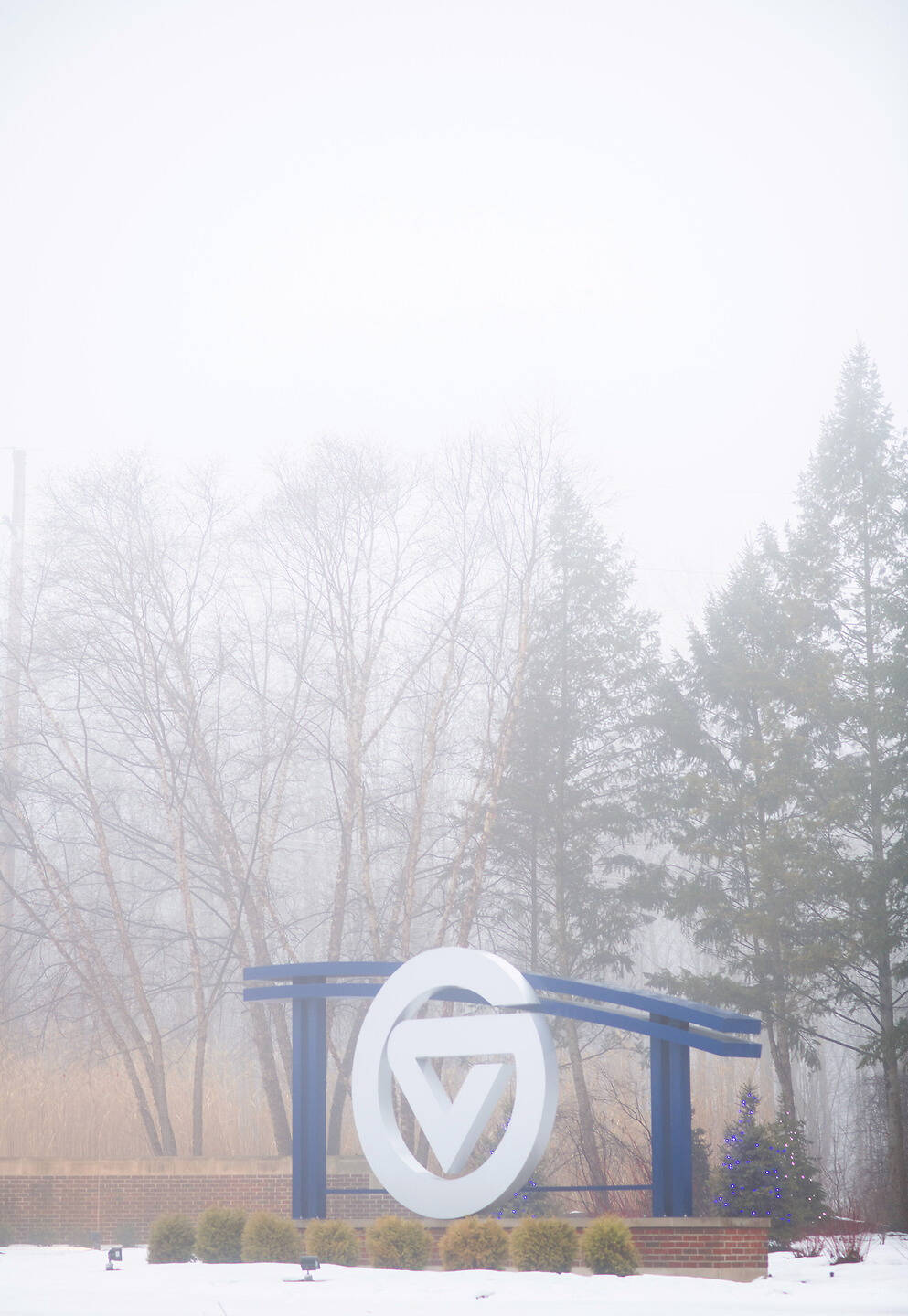 A large white circular logo with a triangular design stands on a blue metal frame in a snowy landscape, surrounded by bare trees and evergreens in heavy fog.
